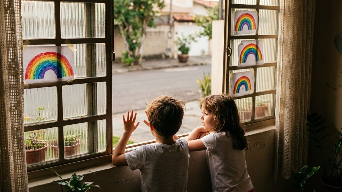 Crianças olhando pela janela de casa a rua vazia, com desenhos de arco-íris colados no vidro