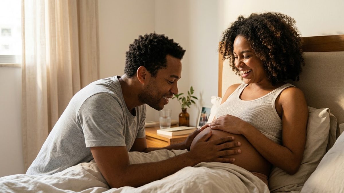 Pai encostando o rosto e a mão na barriga da companheira grávida, falando com o bebê, os dois rindo, no quarto com luz da manhã