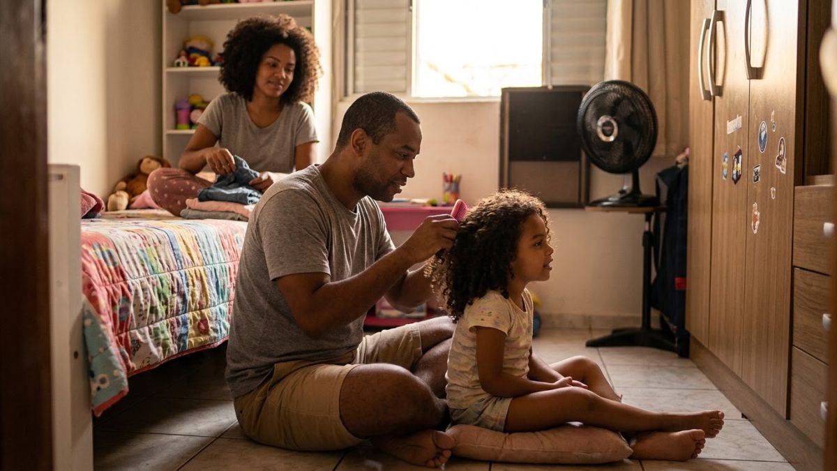 Pai sentado no chão do quarto da filha penteando o cabelo dela com cuidado, os dois conversando, num momento de ternura